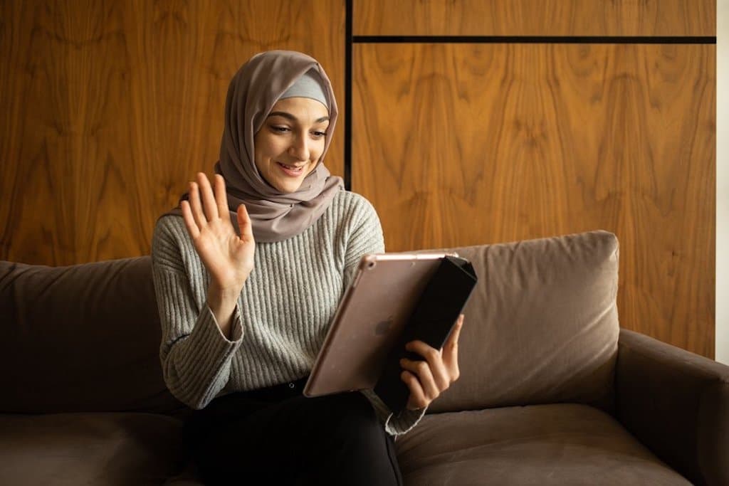 Person with light skin tone wearing a hijab waves hello to someone on their tablet.